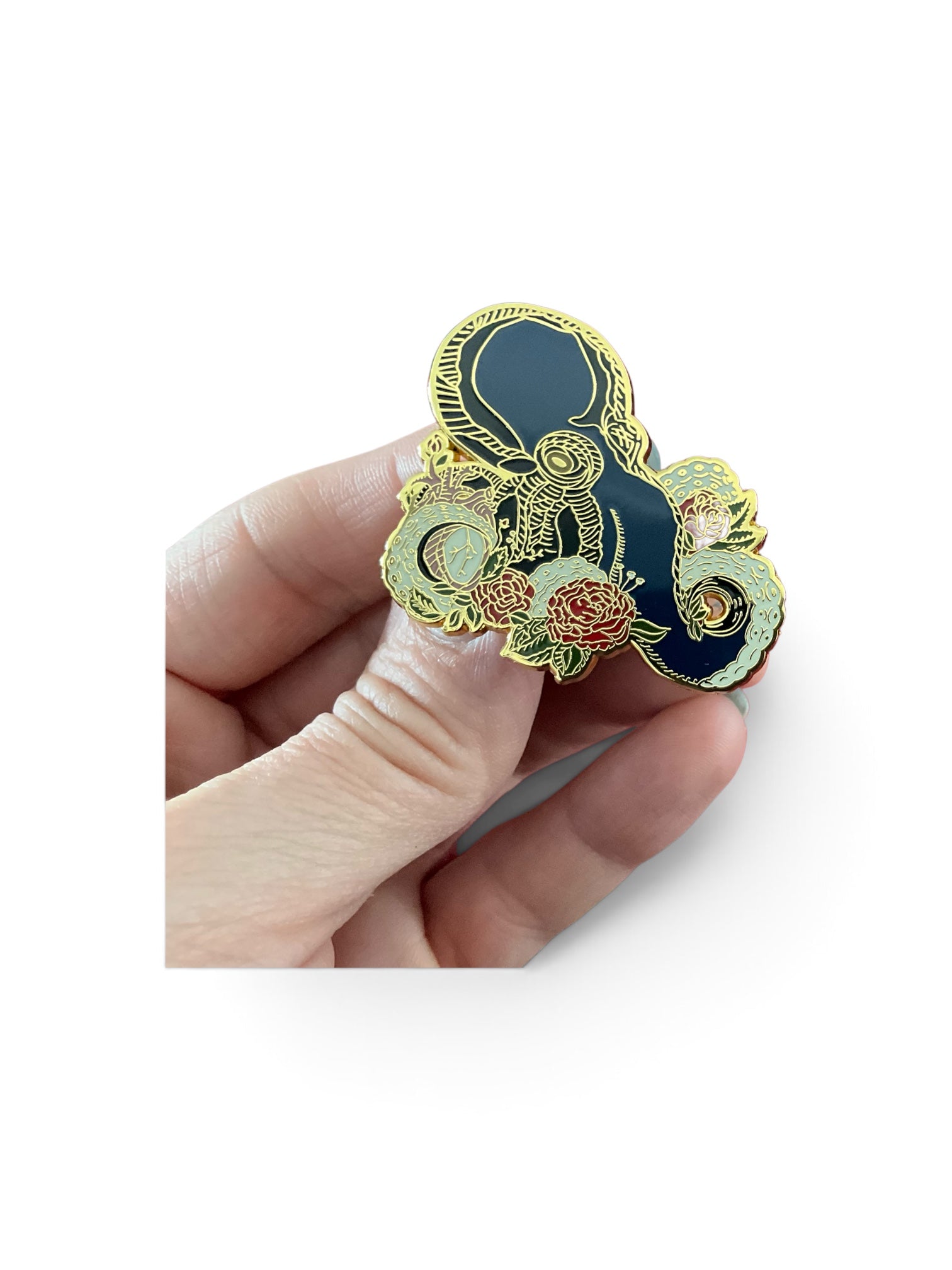 A hand holding a decorative enamel pin shaped like a yin-yang symbol, featuring a dark navy blue center, gold outlines, and floral details in pink, green, and gold, set against a plain white background.