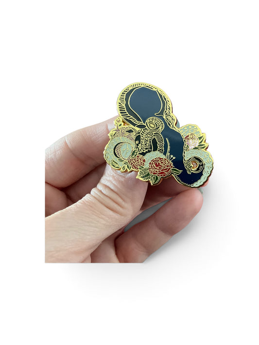 A hand holding a decorative enamel pin shaped like a yin-yang symbol, featuring a dark navy blue center, gold outlines, and floral details in pink, green, and gold, set against a plain white background.